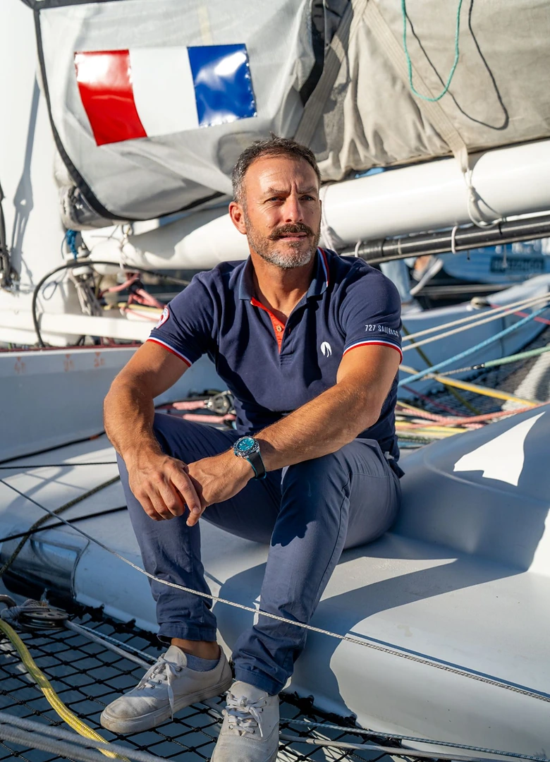 Le skipper Eric Péron assis sur le flotteur de son trimaran, montre Alpina Seastrong Diver Extreme Route du Rhum au poignet